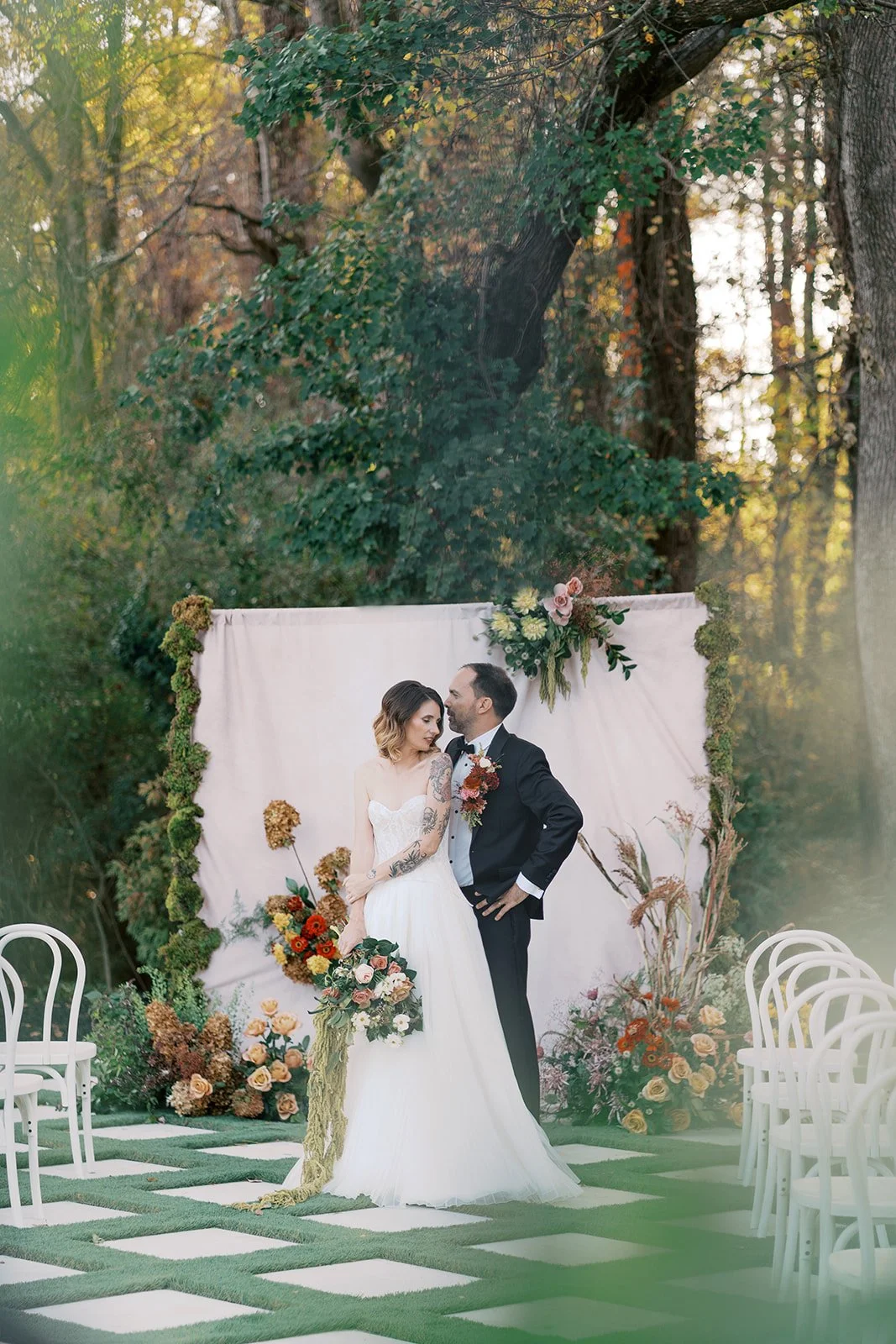 A bride and groom standing close together during their outdoor wedding ceremony, with a floral backdrop and chairs arranged on a grassy checkered pattern.