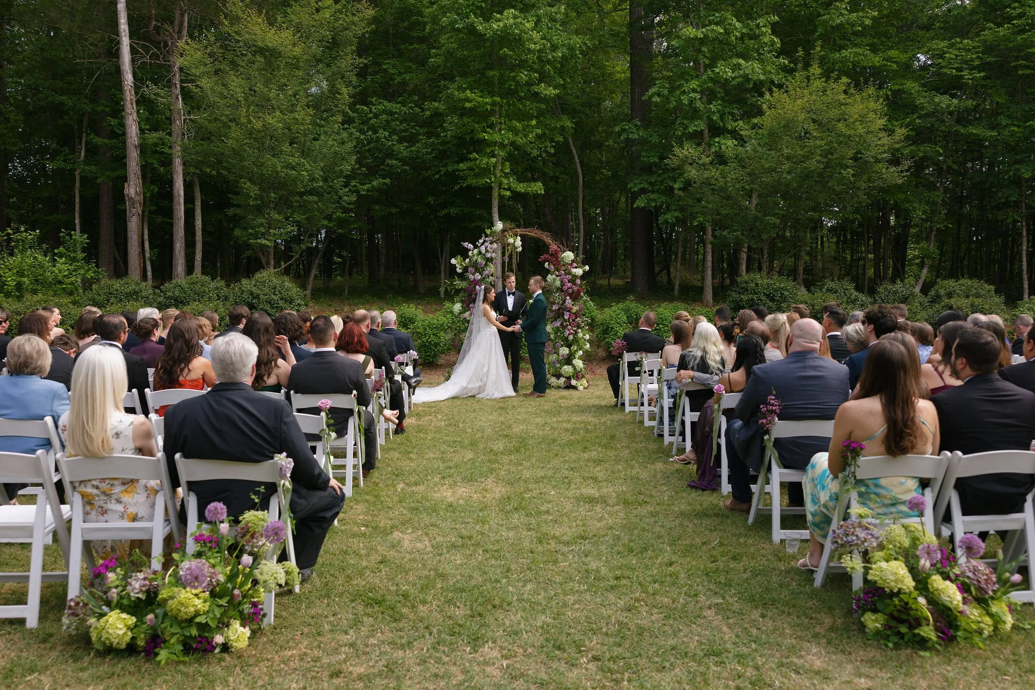 Outdoor garden wedding ceremony with a floral arch and tree line at The Barn of Chapel Hill