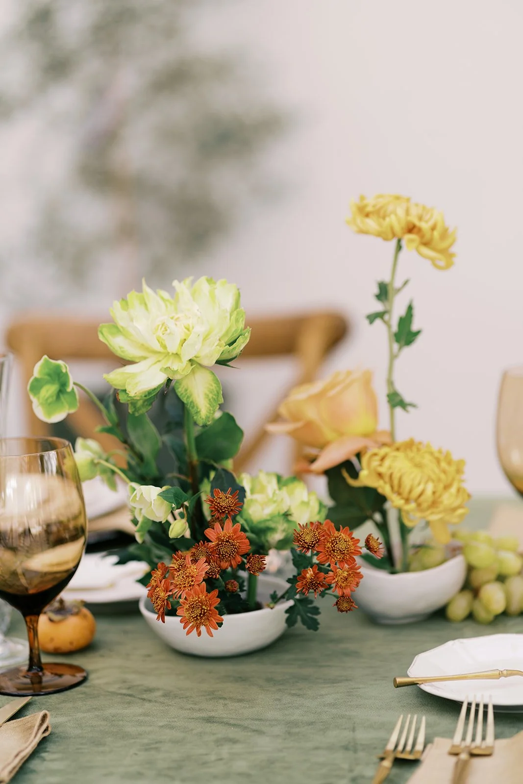 Close-up of a table with a floral centerpiece consisting of yellow, green, and orange flowers, gold-rimmed wine glasses, a white plate, and gold utensils on an olive green tablecloth.