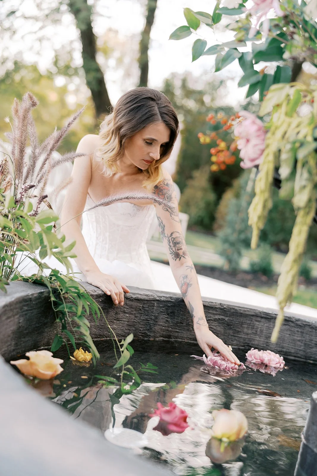 Woman in a wedding dress with tattoos leaning over a water feature with floating flowers and surrounded by greenery and flowers outdoors.