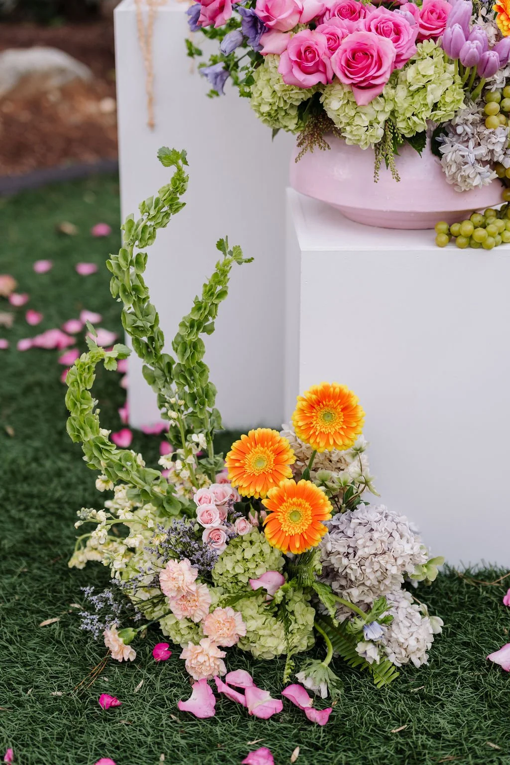 Colorful flower arrangement with pink roses, orange gerbera daisies, purple flowers, and green foliage on a white pedestal outdoors with scattered pink rose petals on grass.