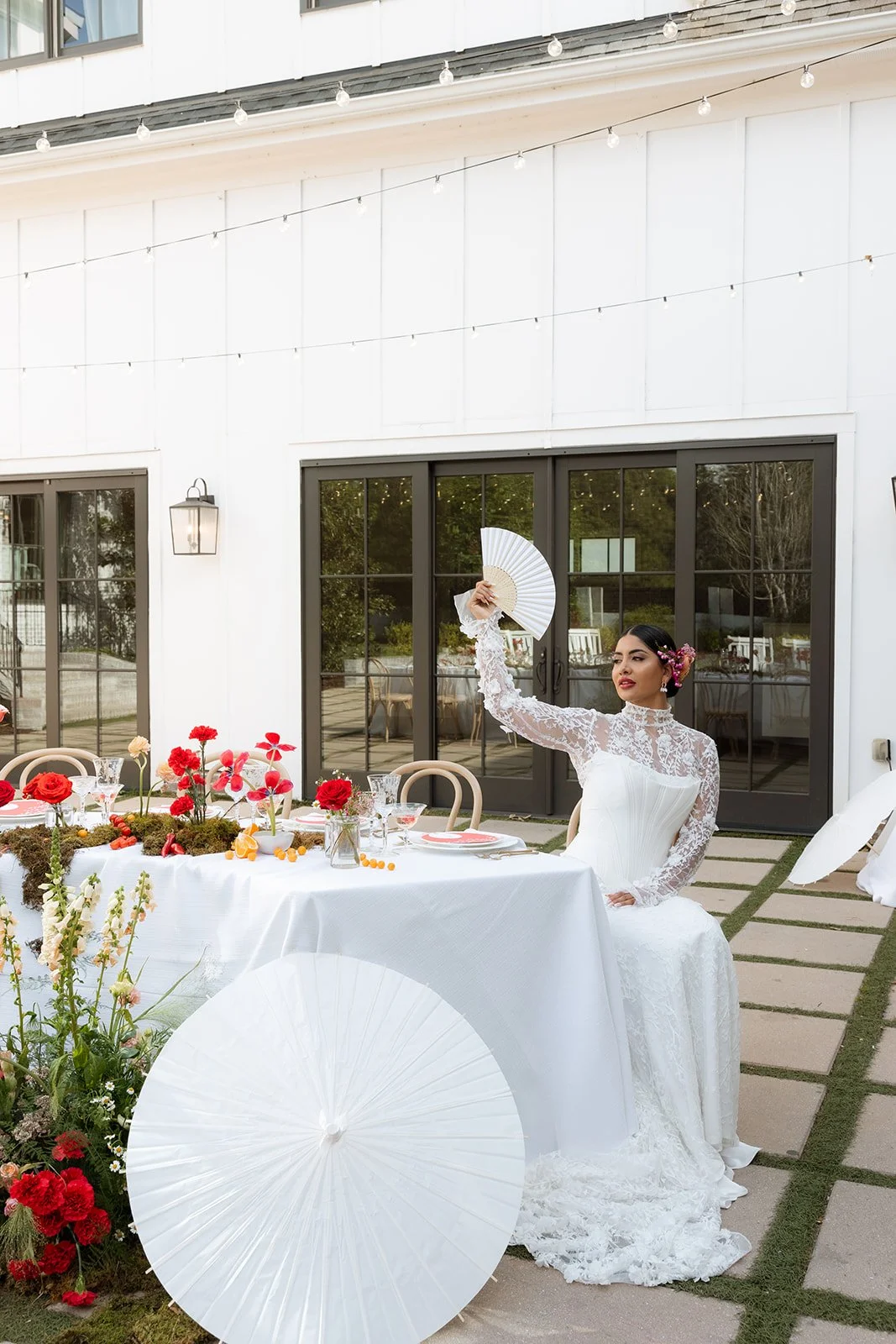 Bride in white lace dress holding a white folding fan at outdoor wedding reception with decorated table and umbrellas.