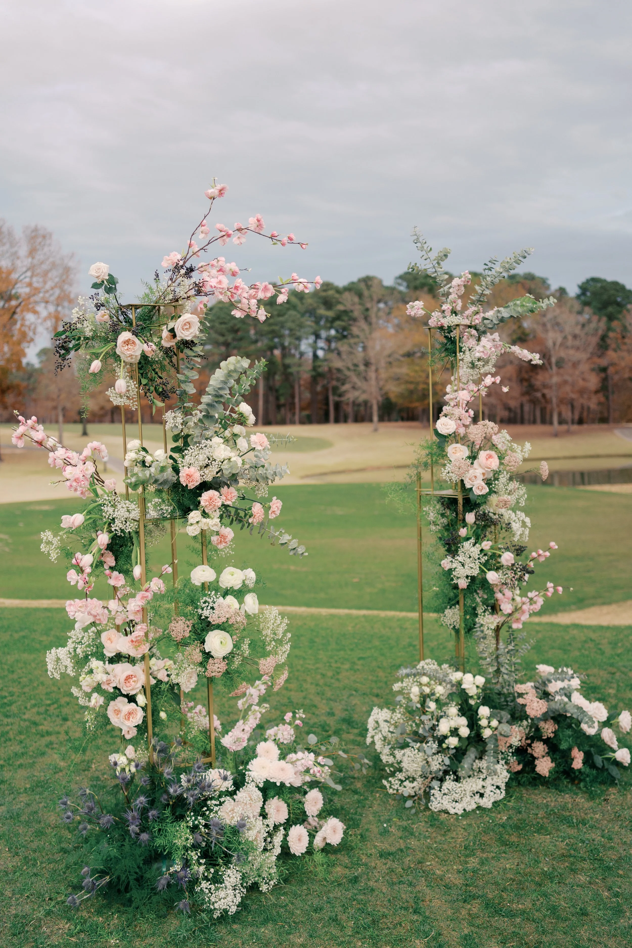 Outdoor wedding altar decorated with pink and white flowers, set against a grassy field and trees with autumn foliage under a cloudy sky.