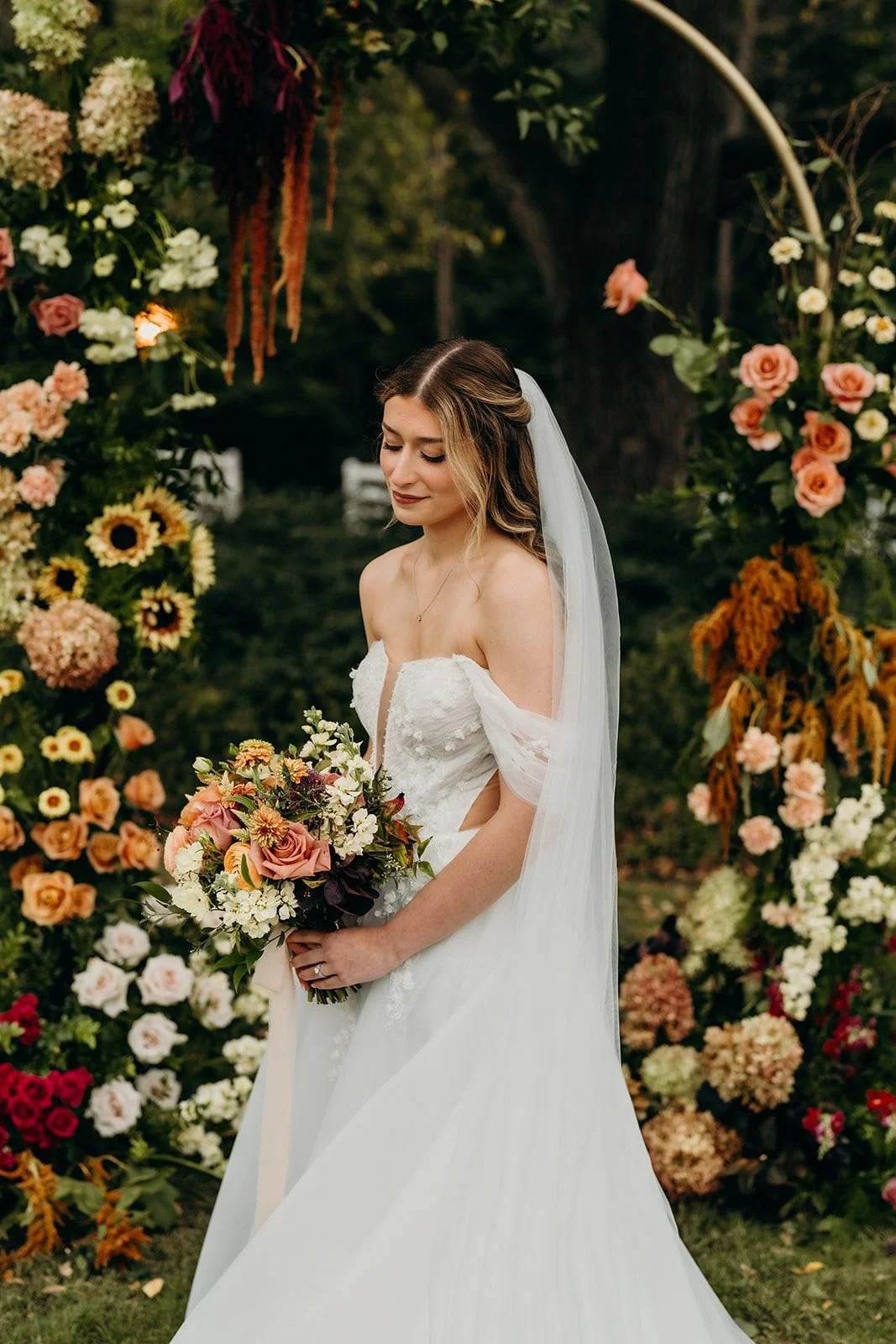 Bride holding a fall bouquet of terracotta roses and ranunculus in front of a floral arch with hydrangea, sunflowers, and hanging amaranthus at The Wakefield Barn near Raleigh