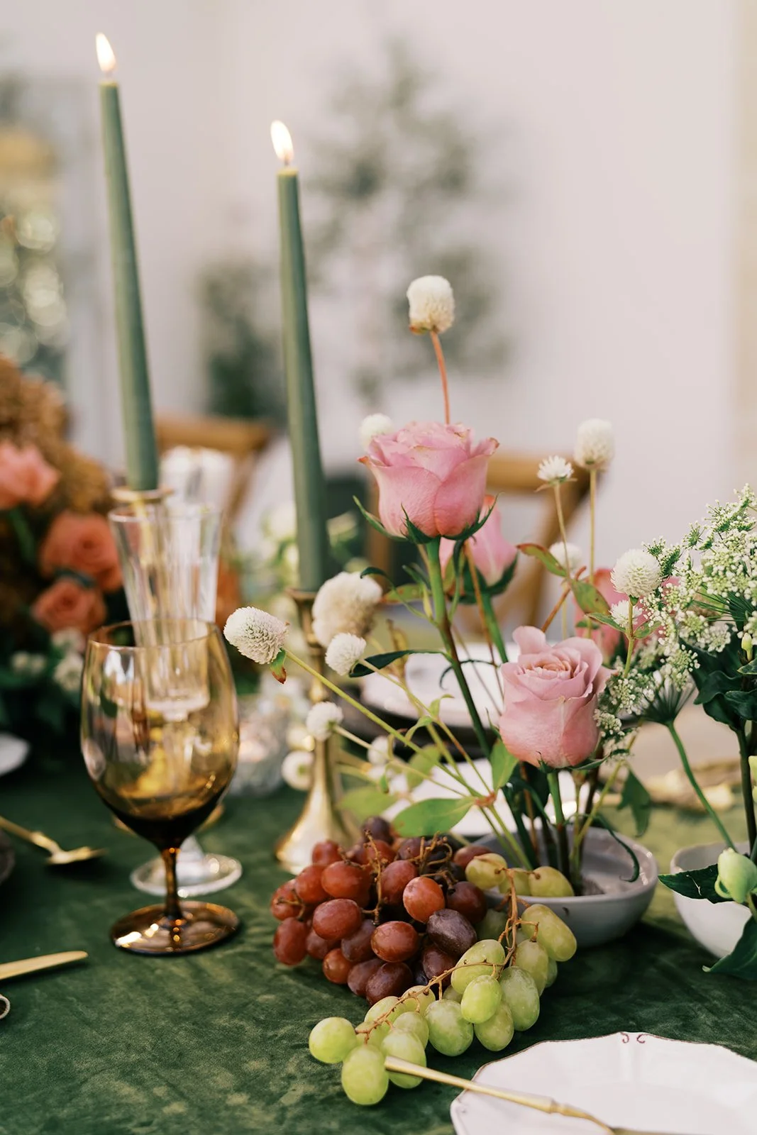 Elegant table centerpiece with pink roses, white flowers, green grapes, and a tall green candle in a glass holder at a formal event.