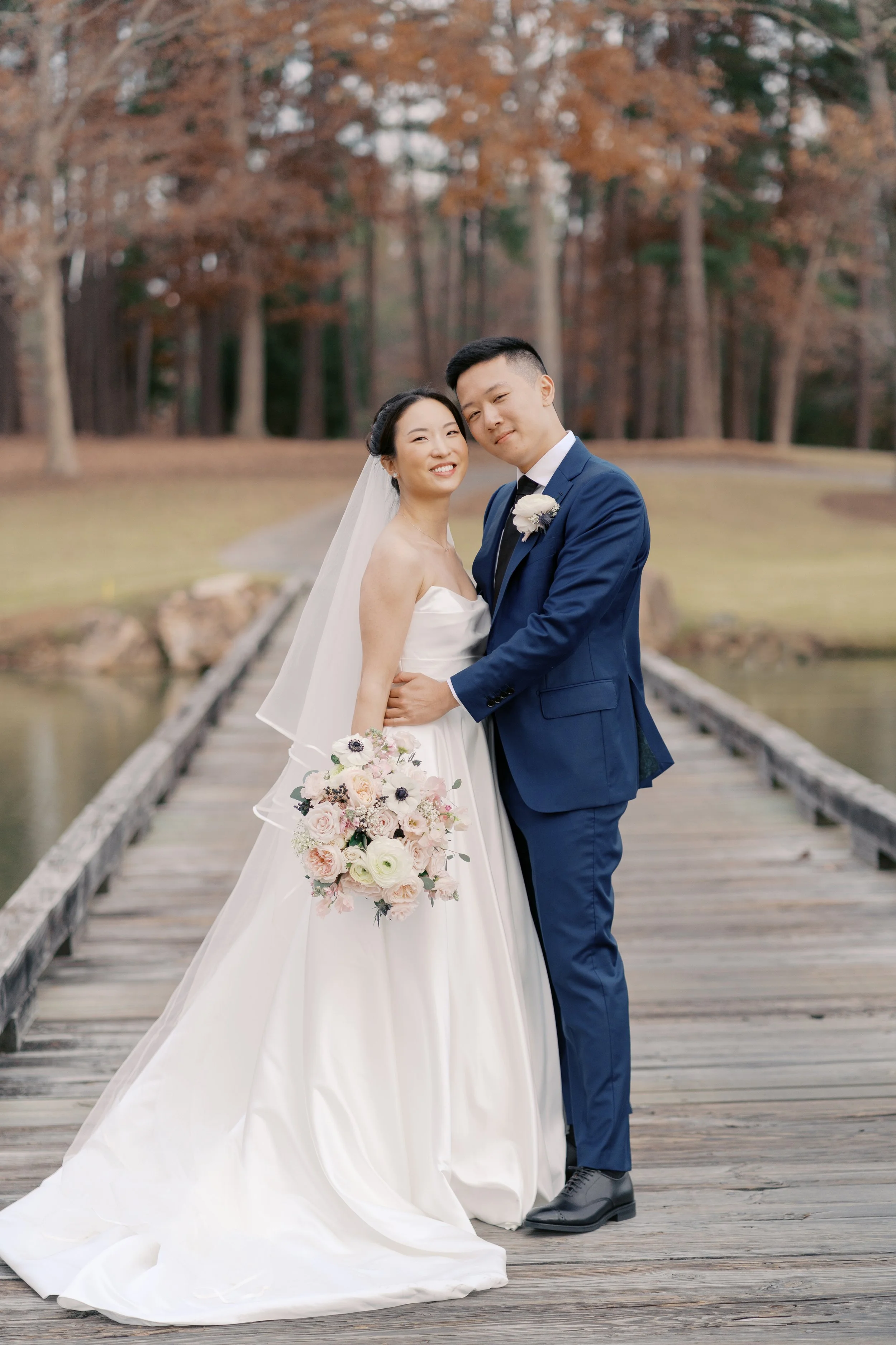 A bride and groom stand on a wooden bridge in an outdoor setting with tall trees and autumn foliage in the background, smiling and posing for a wedding photo.