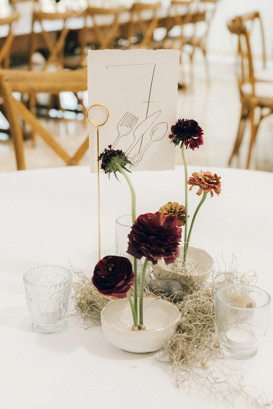 Table centerpiece with dark red and orange flowers in a white bowl, surrounded by glass candle holders and decorative moss, with a napkin featuring utensil sketches standing behind the flowers.
