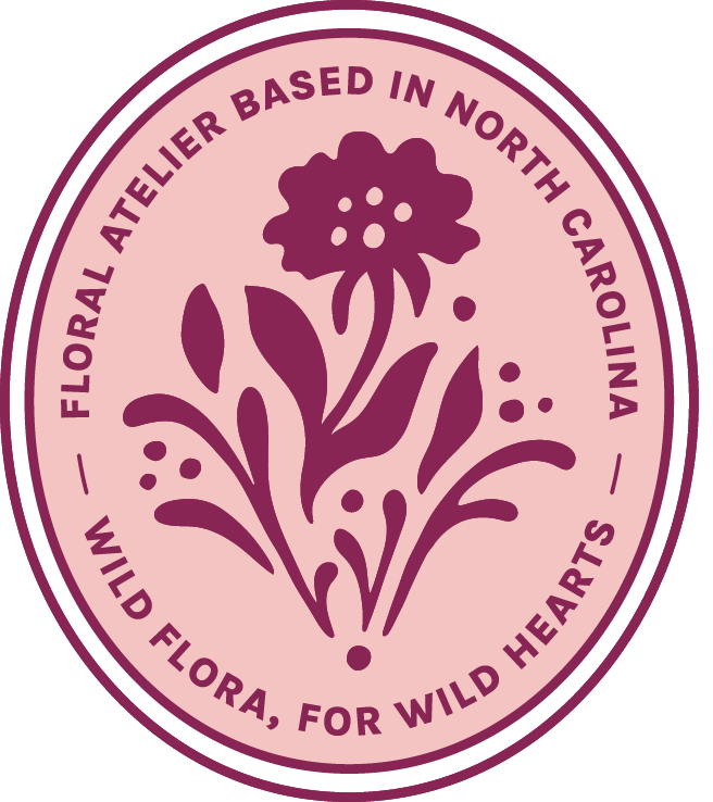 Circular logo with a pink background featuring a purple wildflower illustration and text that reads 'Floral Atelier Based in North Carolina' and 'Wild Flora, For Wild Hearts'.