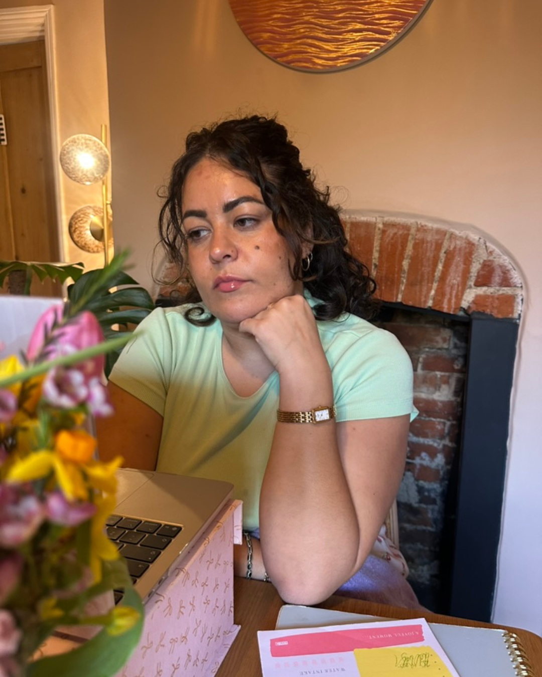 A woman with dark curly hair and light skin rests her chin on her hand, sitting at a table with a laptop and a flower arrangement, in a room with a brick fireplace, warm lighting, and wall decorations.