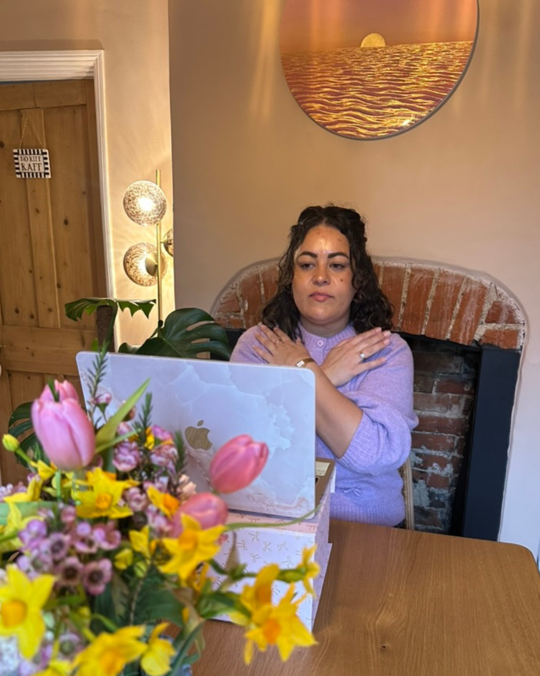 A woman in a lavender sweater sitting at a wooden table with a laptop in front of her, crossing her arms over her shoulders, with a large bouquet of pink and yellow flowers in the foreground and a brick fireplace behind her.