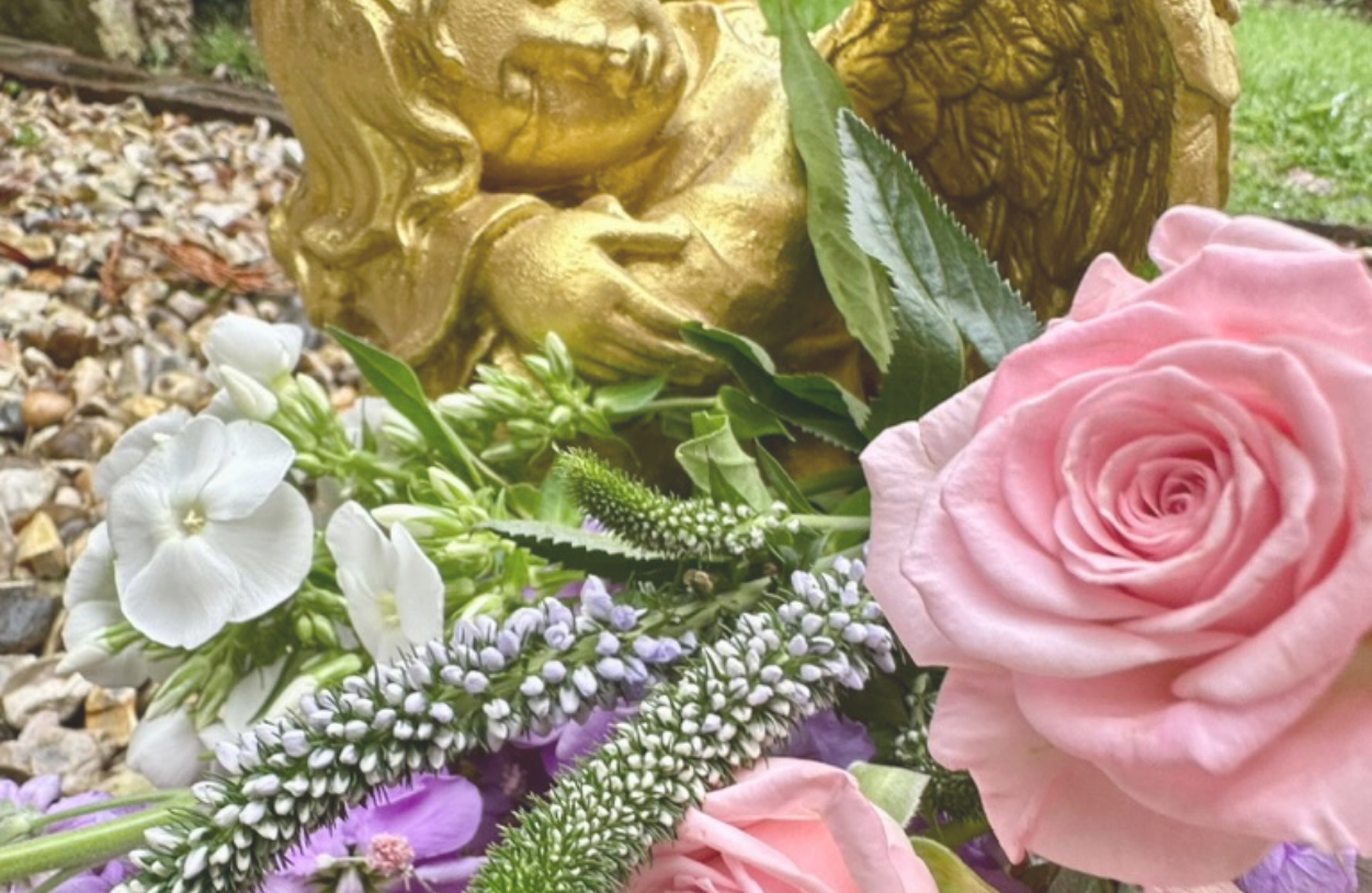 Close-up of various flowers including pink roses, white blooms, and purple flowers, with a golden statue of a face in the background.