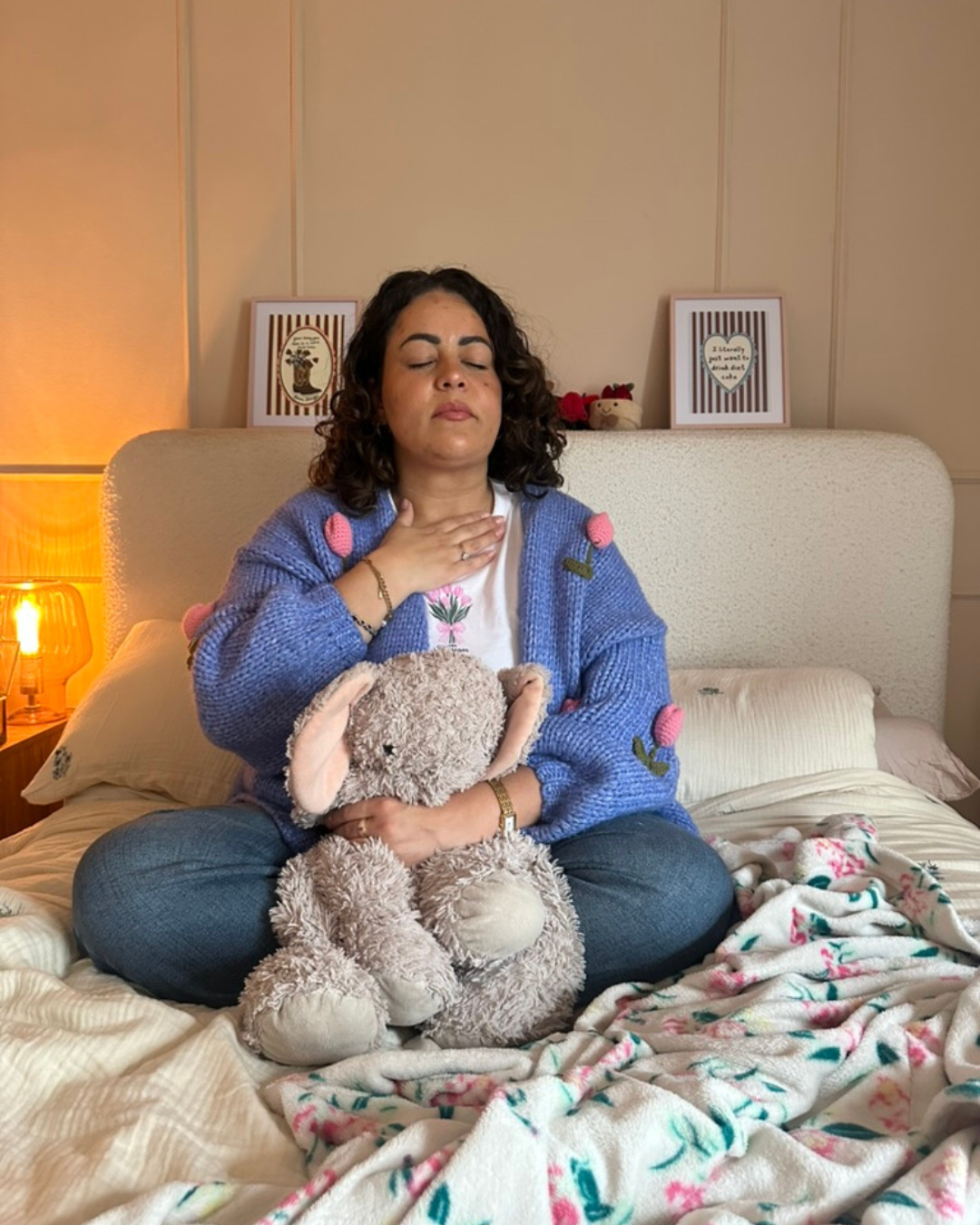 A woman sitting cross-legged on a bed, holding a plush bunny toy, with her eyes closed and hand on her throat, appearing to meditate or relax in a cozy bedroom.