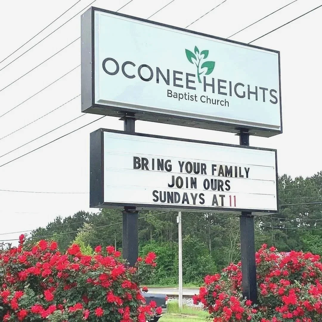 Sign for Oconee Heights Baptist Church with a message encouraging families to join on Sundays at 11, surrounded by red flowers.