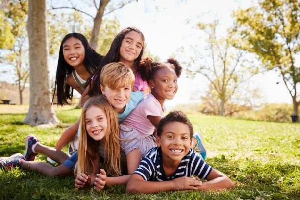 Group of six diverse children lying on the grass in a park, smiling and enjoying a sunny day.