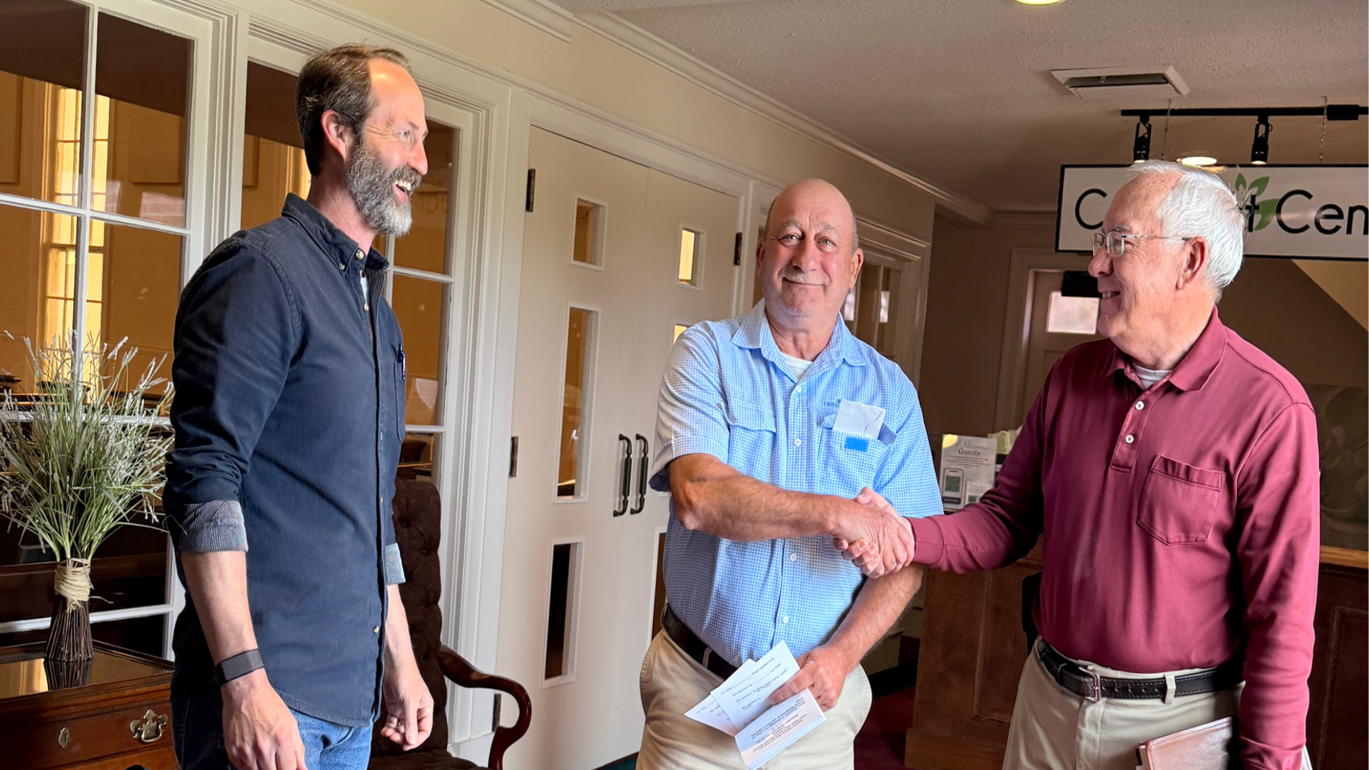 Three men inside a building, two of them shaking hands, appear to be having a conversation, with a woman on the left observing. The background shows a floral arrangement and a sign partially visible with the words 'C...C'.