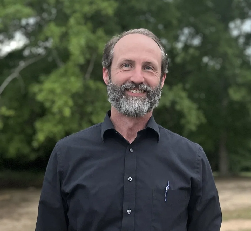 A smiling man with a beard and short hair, wearing a black button-up shirt, standing outdoors with green trees in the background.