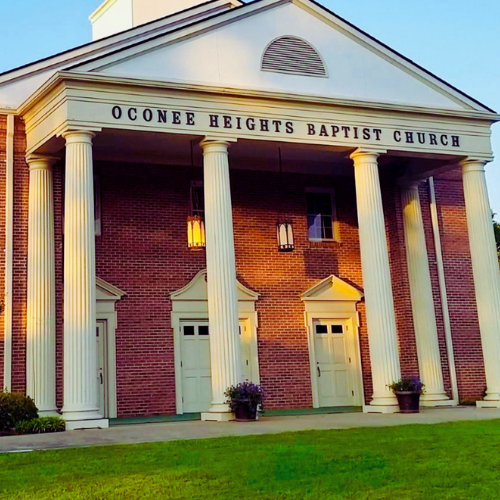 Front view of Oconee Heights Baptist Church with brick facade, white columns, and three doors, taken during sunset.