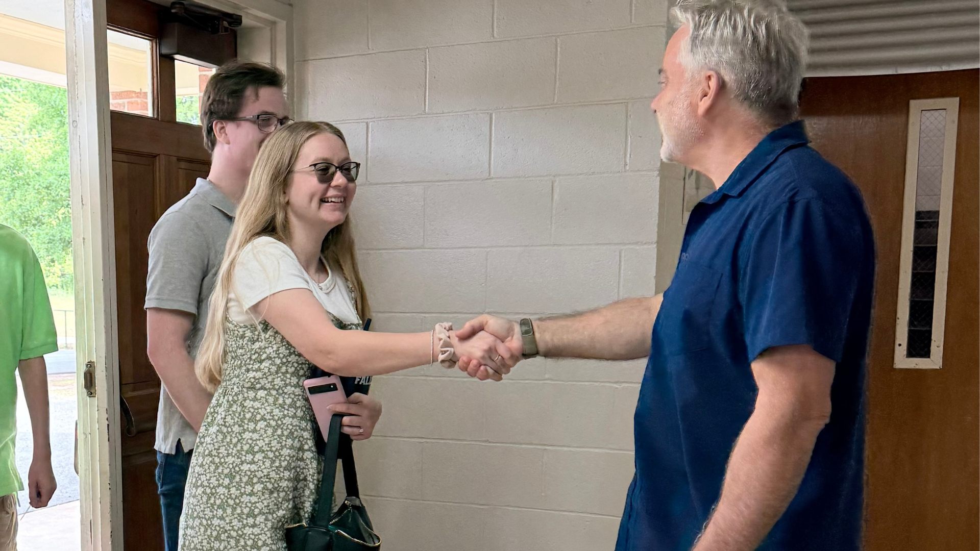 A young woman with long blonde hair and glasses shaking hands with an older man with gray hair and a beard inside a building. Two other people are partially visible in the background, standing near a door with a window and a selector switch. The woman is smiling and holding a phone and a bag.