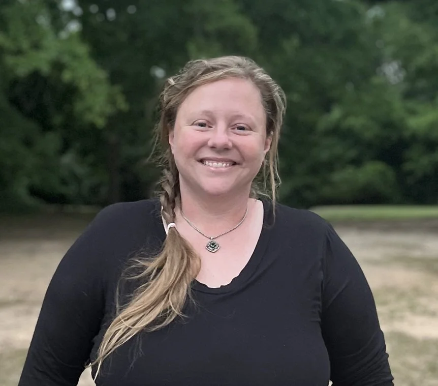 A woman with light skin, long braided hair, wearing a black top and necklace, smiling outdoors with trees in the background.