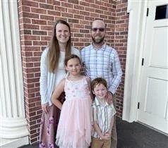 A family of four standing outside a brick house, smiling. The woman has long hair and is wearing a white top. The man has a beard and sunglasses, wearing a plaid shirt. The two children, a girl in a pink dress and a boy in a striped shirt, stand in front of them.