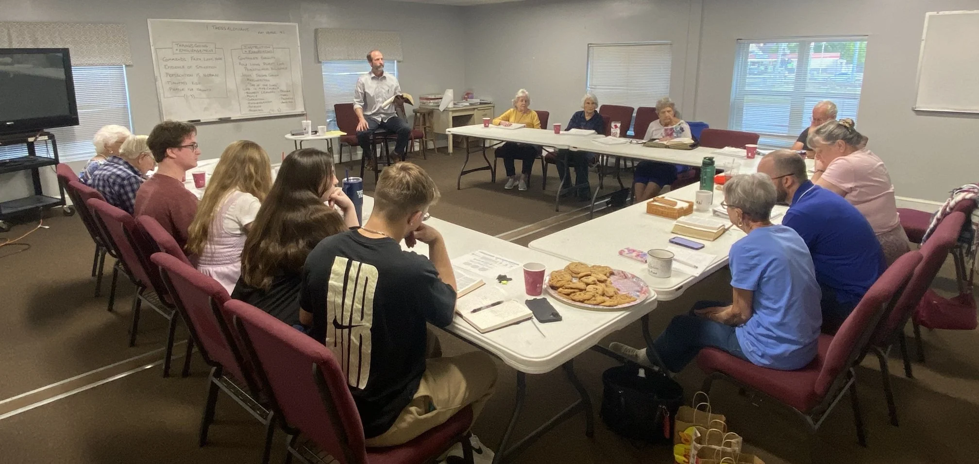 A group of people sitting around a U-shaped conference table in a meeting room, with a man standing at the front holding a book. The table has papers, notebooks, cups, and a large plate of cookies.