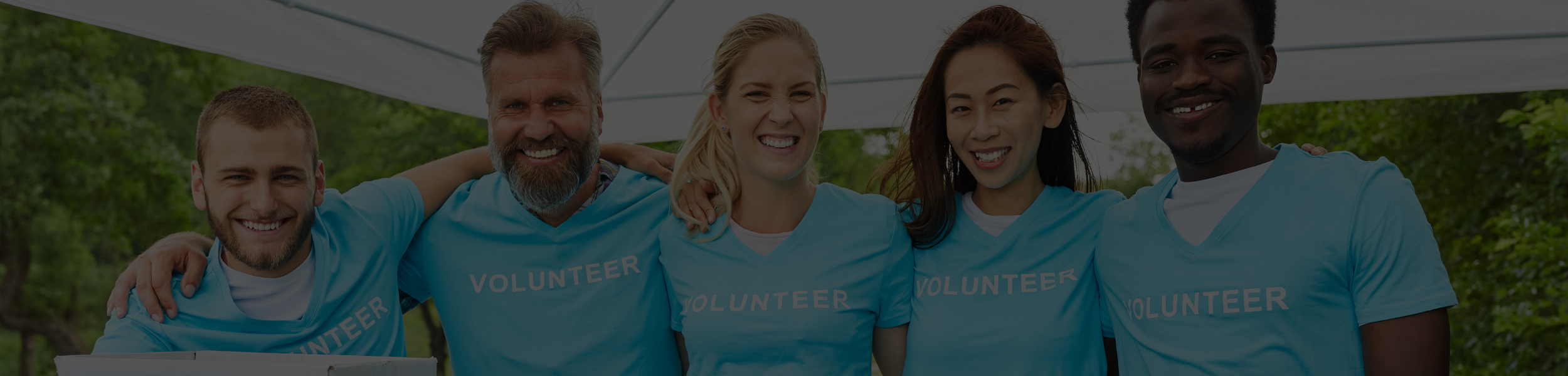Group of five volunteers smiling and hugging outdoors, wearing matching light blue shirts with 'VOLUNTEER' printed on them.
