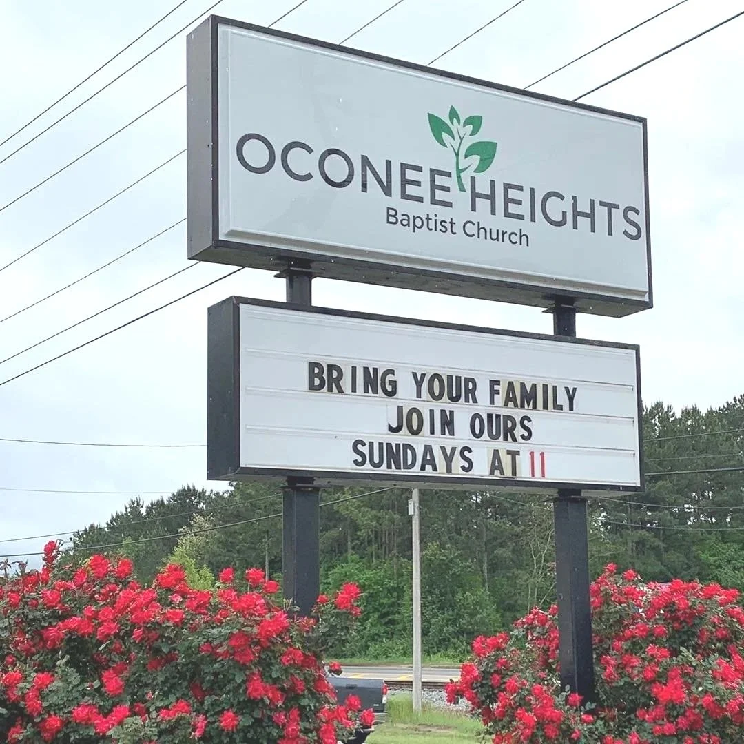 Sign for Oconee Heights Baptist Church with an announcement to bring family and join on Sundays at 11, surrounded by pink flowers.