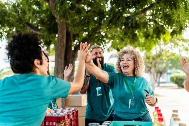 A group of people in turquoise shirts giving each other high-fives outdoors under a tree, smiling and celebrating.