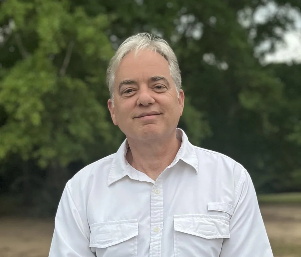 A middle-aged man with gray hair, dressed in a white button-up shirt, standing outdoors in front of green trees.