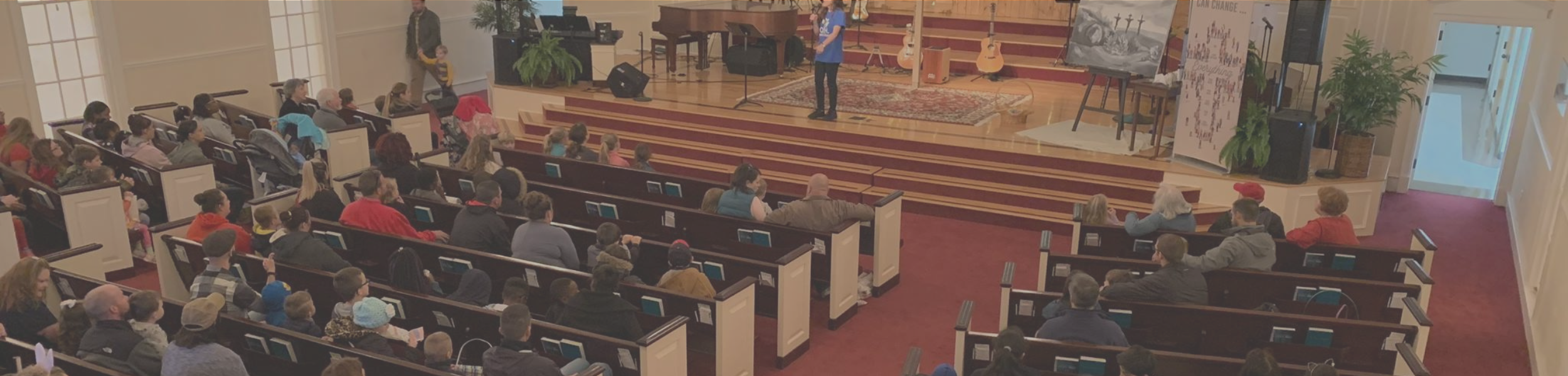 A church service or event with a congregation seated in pews facing an elevated stage. The stage has musical instruments, including guitars and a piano, and a speaker is speaking or singing into a microphone. The room is decorated with plants and artwork, and a large painting or poster is visible on the right side.