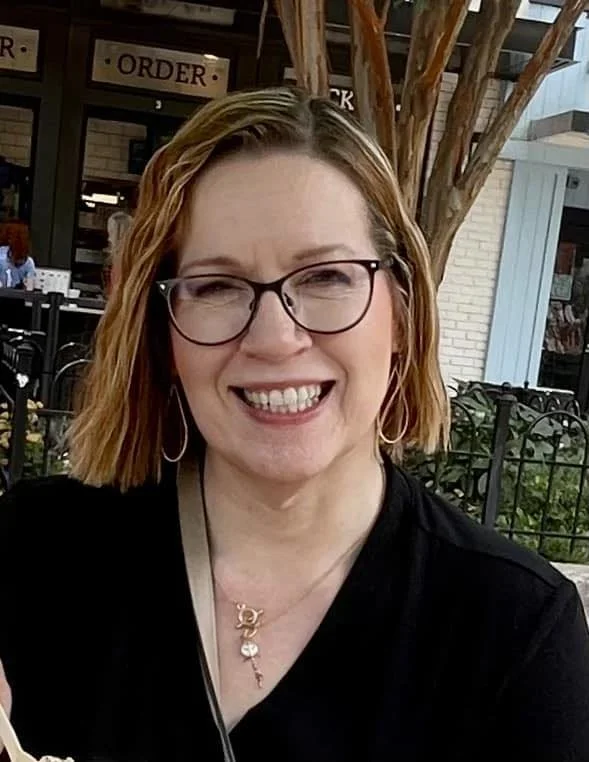 A woman with shoulder-length red hair, glasses, and hoop earrings smiling at the camera. She is wearing a black top and is outdoors near a restaurant with a tree and a sign that says 'ORDER' visible in the background.