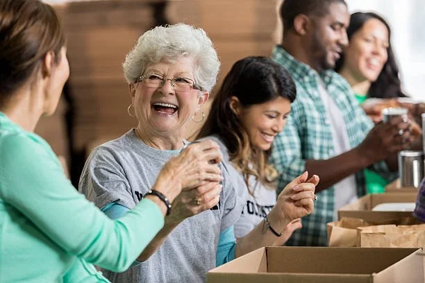 Smiling diverse group of people working together, packing brown paper bags at a table in a bright room.