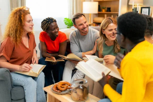 Five people sitting on a couch, engaging in a lively discussion with books in hand, in a cozy living room setting.