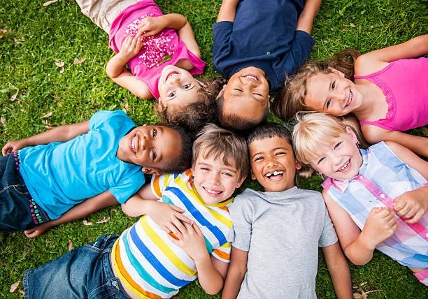 Six children lying on grass in a circle, smiling and looking up at the camera.