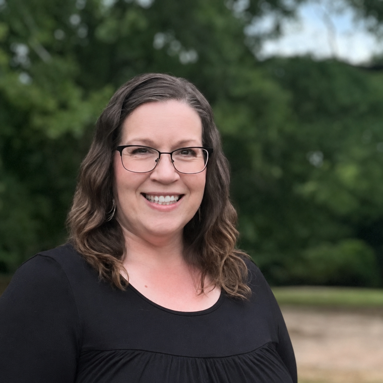 A woman smiling outdoors with trees in the background.