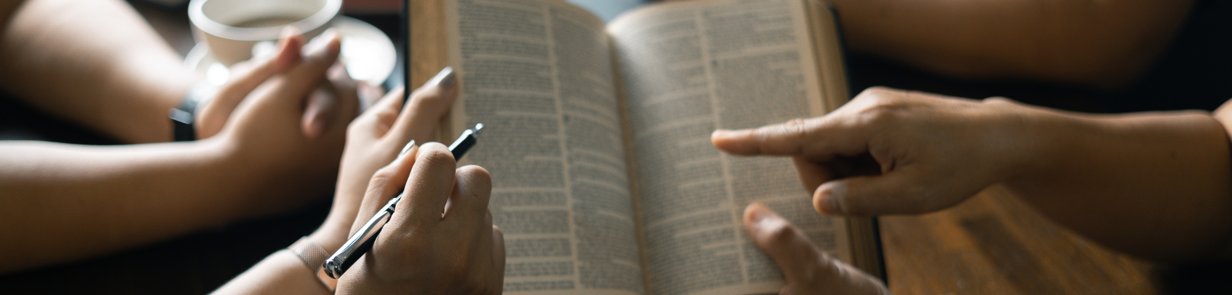 Two people reading a book together, one holding a pen, the other with a hand pointing to the pages.