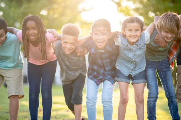 A diverse group of children standing outdoors with arms around each other, smiling and laughing in a park during daytime.
