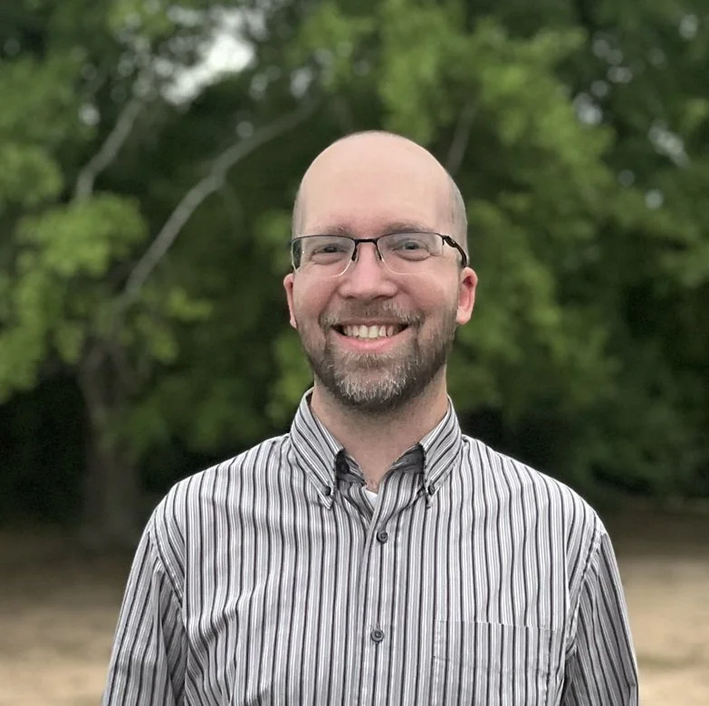 A smiling man with glasses and a beard, wearing a striped shirt, standing outdoors with green trees in the background.