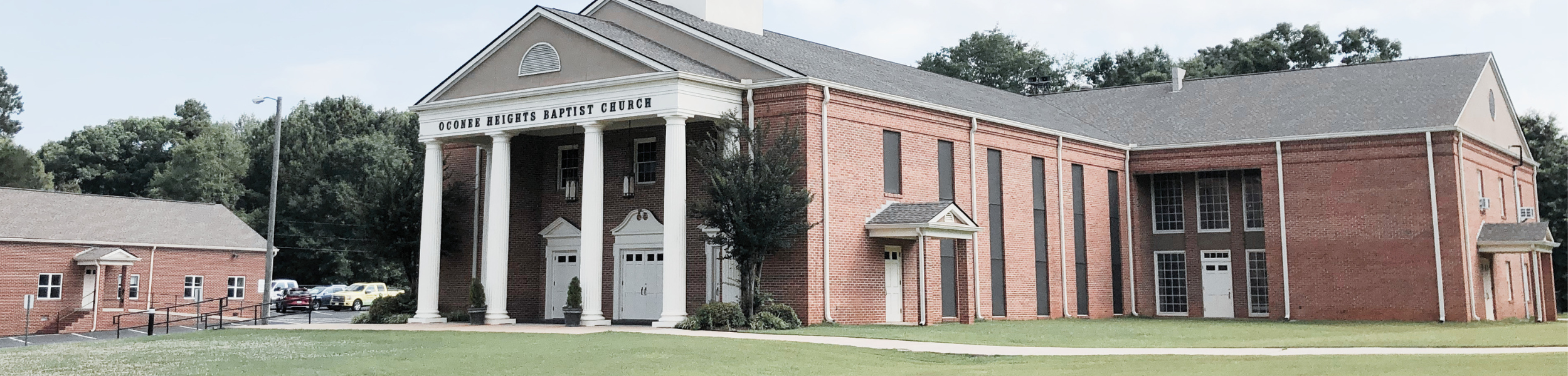 Front view of Oconee Heights Baptist Church building with brick exterior, white columns, and a grassy area in front.