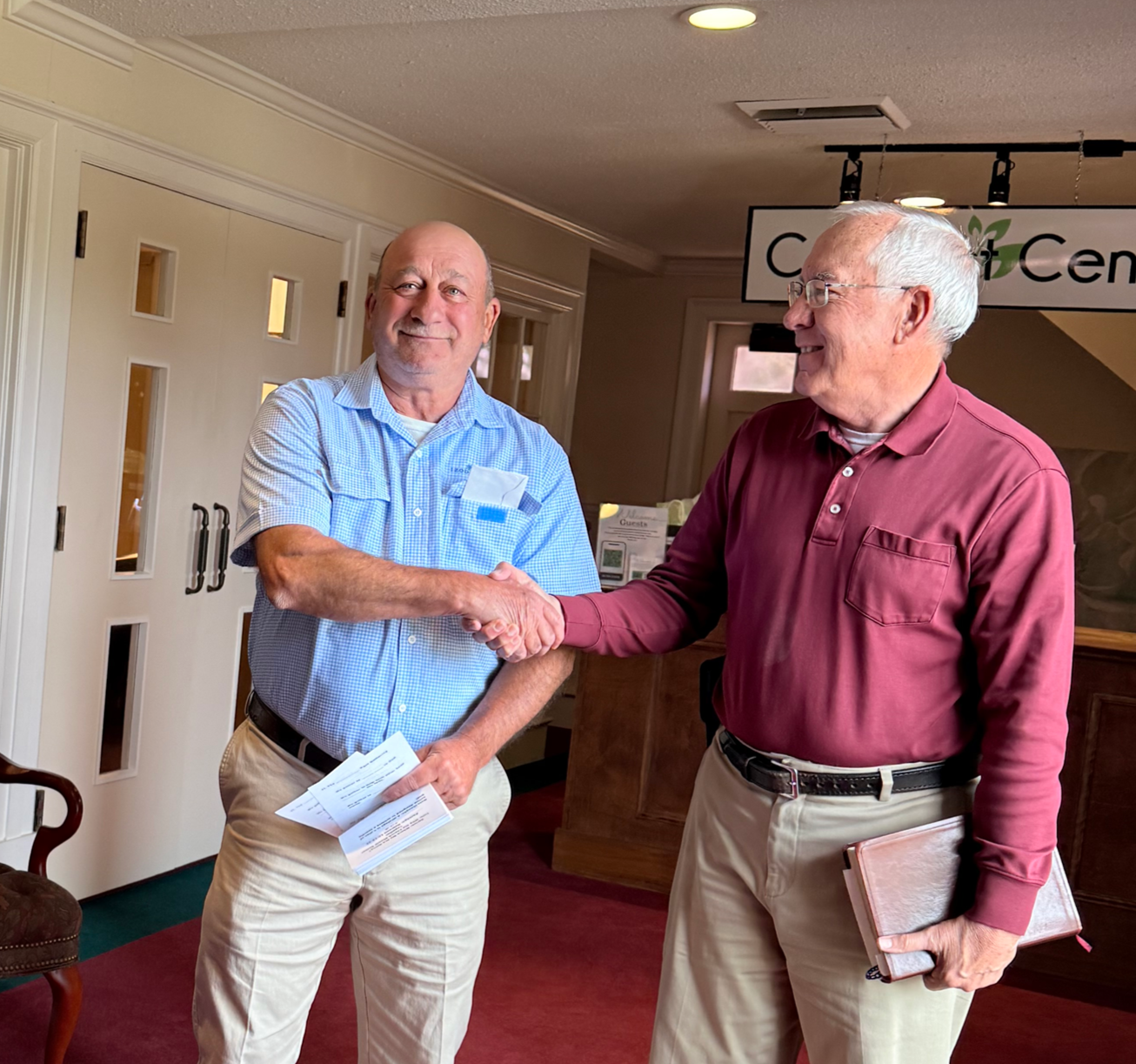 Two older men shaking hands indoors, one wearing a blue checkered shirt and the other in a maroon shirt, with papers in hand, smiling.