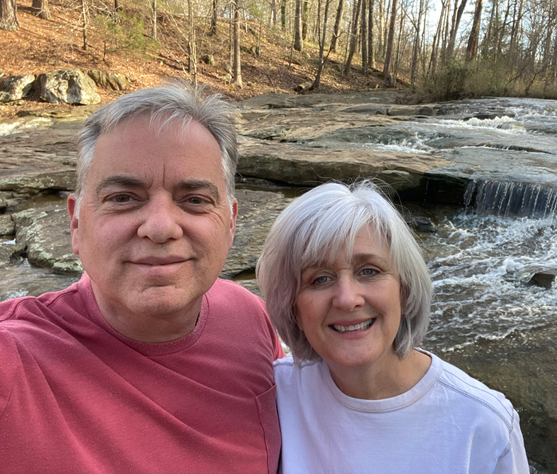 A smiling man and woman taking a selfie outdoors near a flowing creek with rocks and trees in the background.