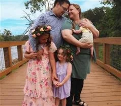 Family of four standing on a wooden bridge outdoors, smiling, with two young girls and two adults, one adult holding a pregnant belly