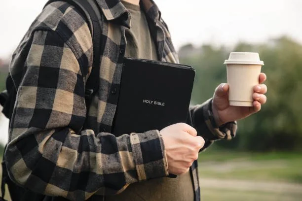 Person holding a disposable coffee cup and a Holy Bible outdoors, wearing a plaid shirt and backpack.