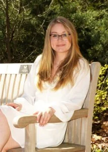 Young woman with glasses sitting on a wooden bench outdoors, surrounded by greenery, smiling.