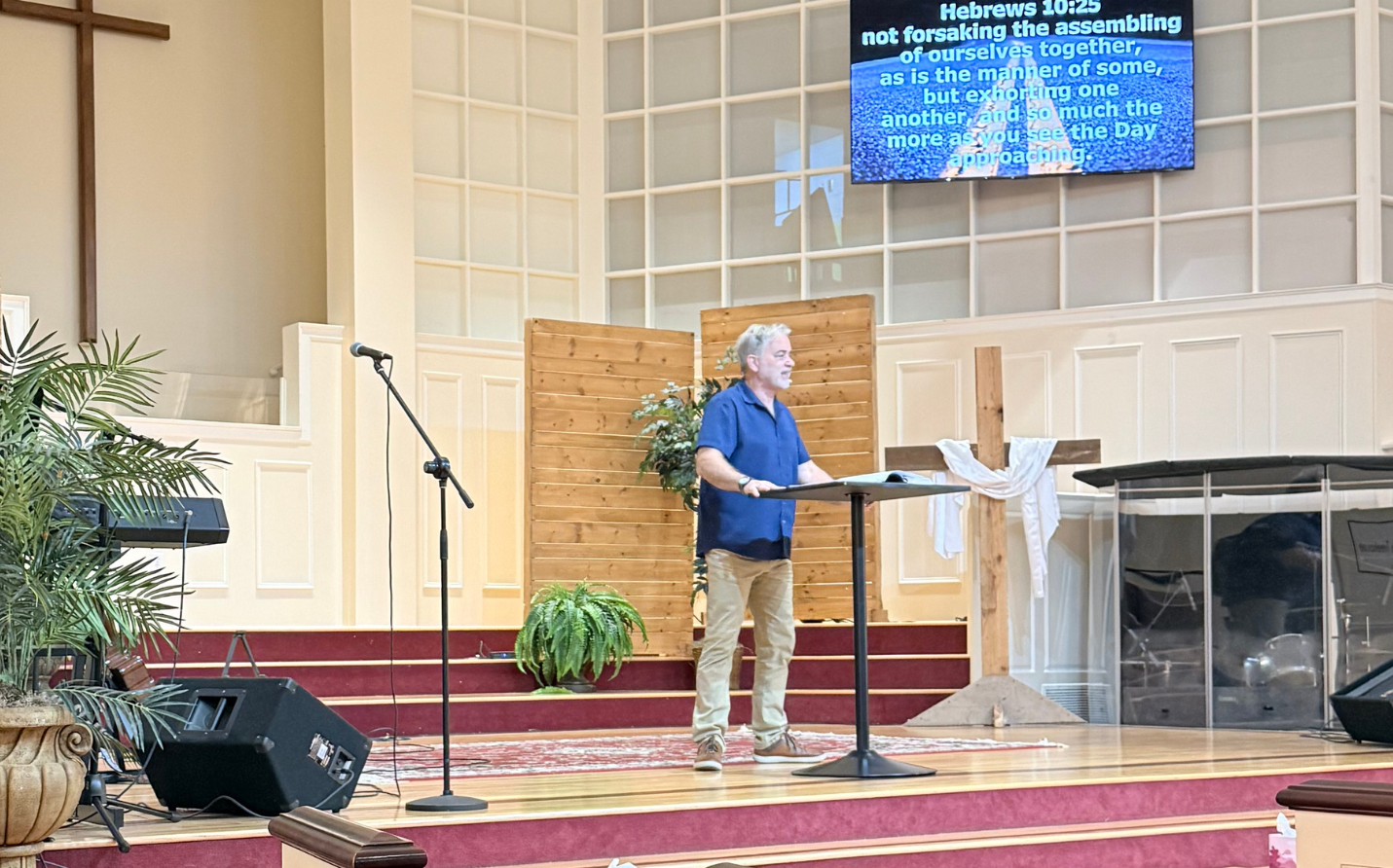 A man standing at a lectern speaking in a church, with a cross and plants nearby, a microphone on a stand, and a digital screen displaying Bible scripture in the background.