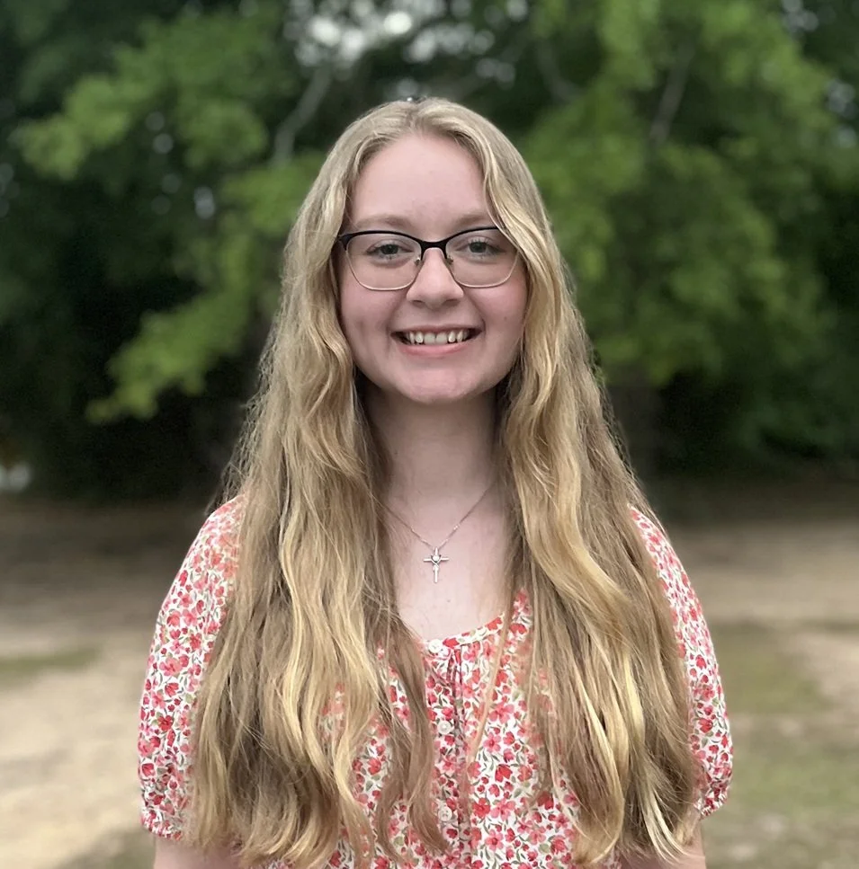 A young woman with long, wavy blonde hair and glasses, smiling outdoors with a background of green trees and a dirt area.