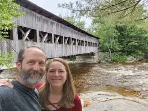 A smiling couple taking a selfie in front of a covered wooden bridge over a river with lush green trees around.