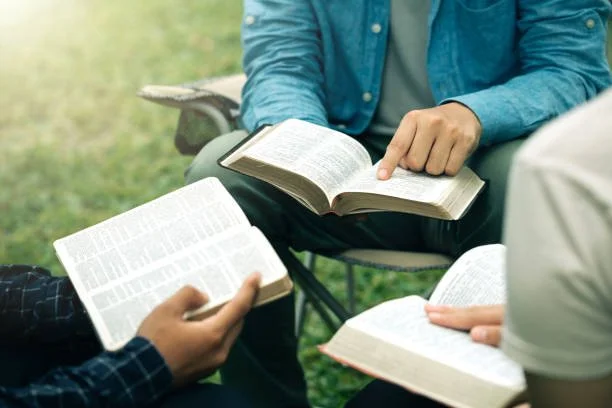 Three people sitting outdoors on the grass, each holding an open Bible and reading.