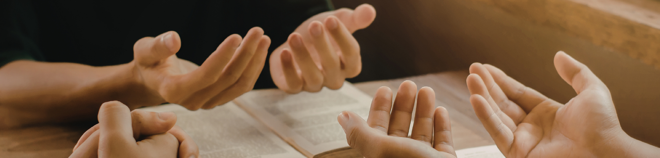 Four hands are shown over an open book, with two of the hands palm-up and two of the hands mid-gesture as if in conversation or explanation.