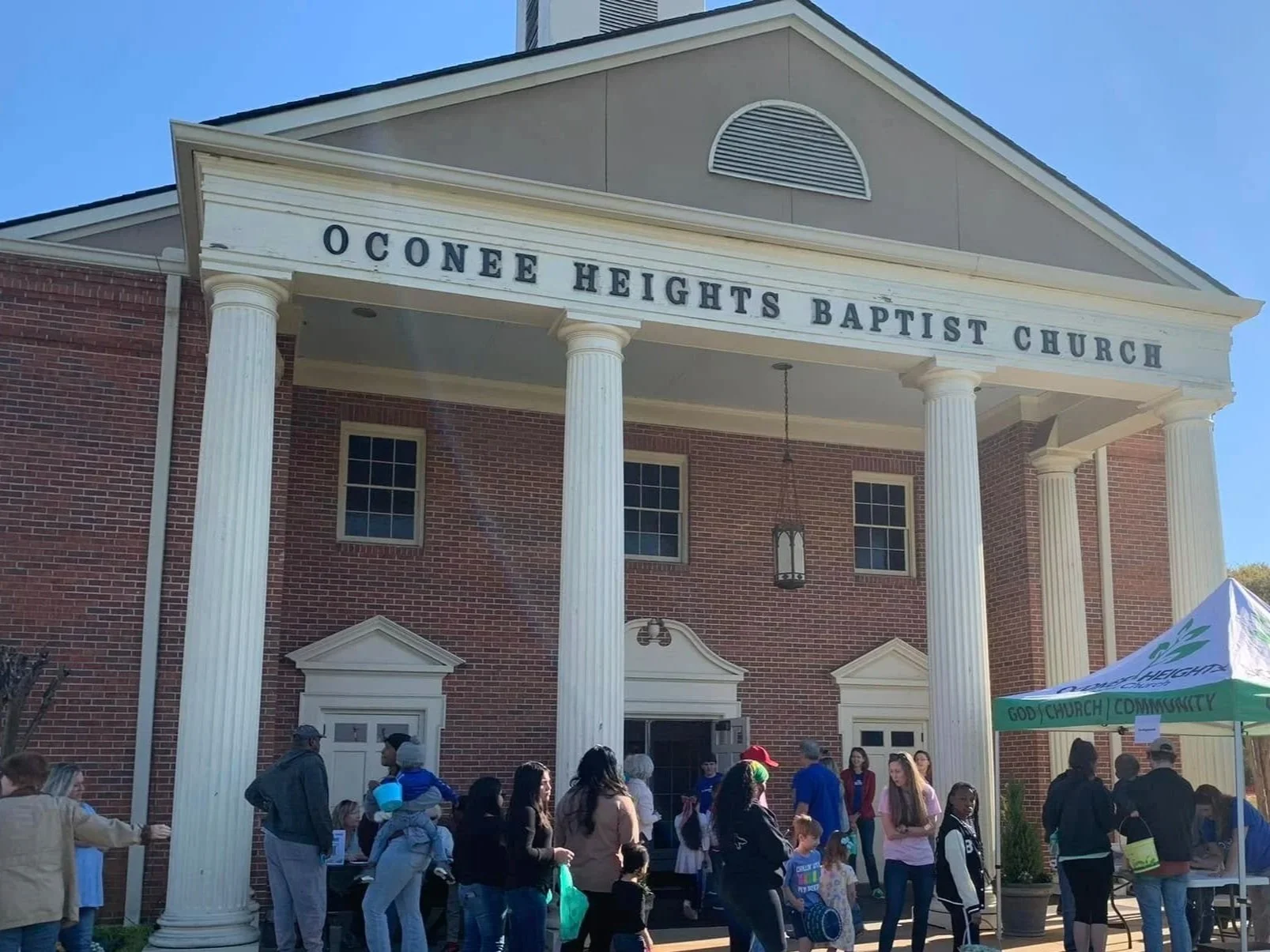 People gathered outside Oconee Heights Baptist Church, a brick building with white columns and a sign above the entrance.